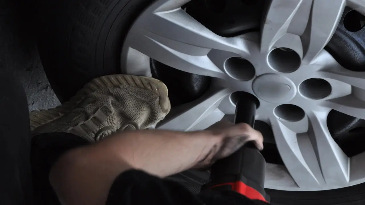A hand running an impact wrench on a car wheel's lug nuts, the tire beside it; the wheel's centre badge blurred.