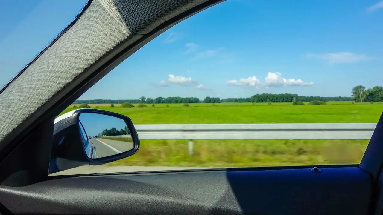 A view from a moving car's passenger window: the door mirror, a guardrail and green fields; window markings blurred.