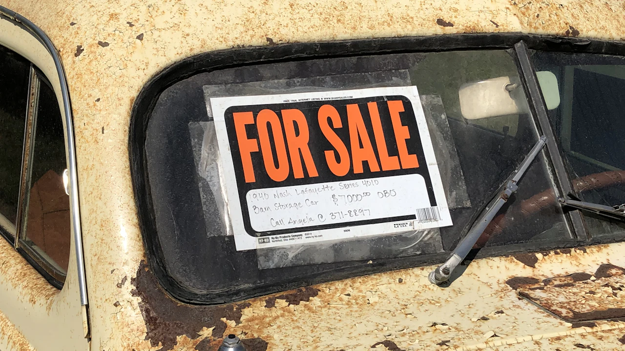 A hand-lettered FOR SALE card taped inside the windscreen of a rusting cream-coloured vintage car, the asking price written on it in biro