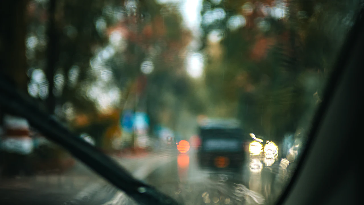 The view from a driver's seat through a rain-spotted windscreen, with a wiper arm and blade crossing the lower left of the glass, the dark A-pillar on the right, and out of focus ahead a wet road with the red tail lights of a car and the headlights of oncoming traffic under trees.
