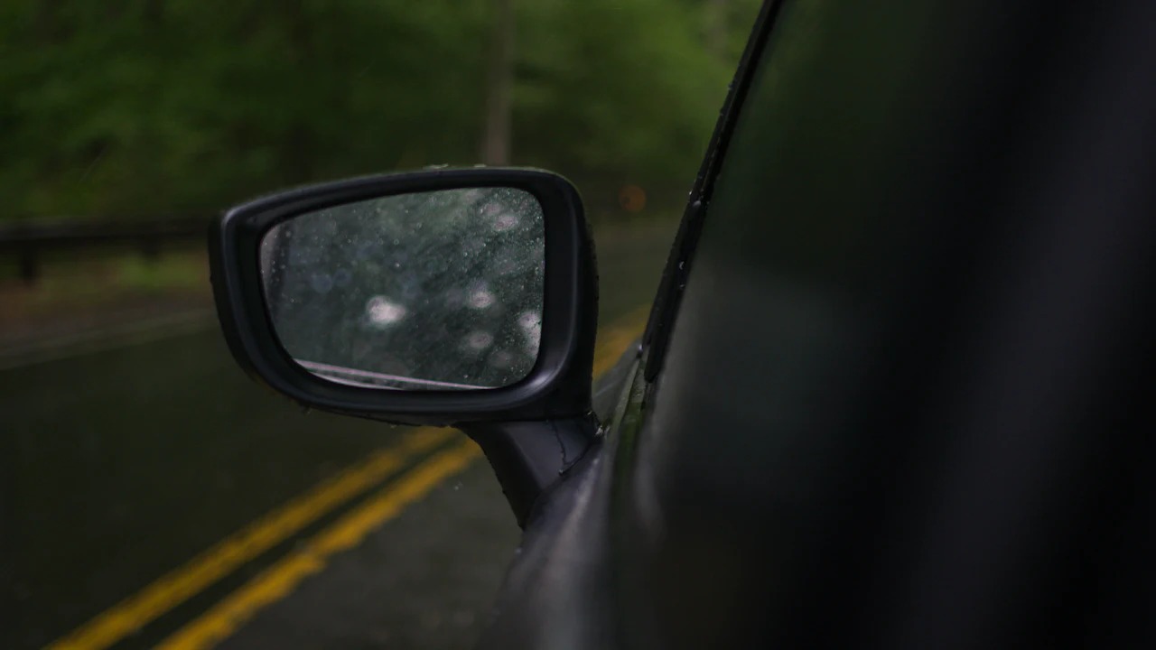 A car wing mirror speckled with rain on a wet tree-lined road, double yellow lines behind