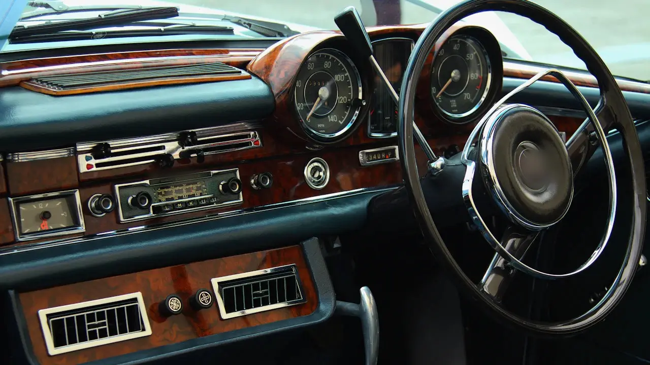 The wood-trimmed dashboard of a classic car, with round gauges, a chrome radio and a thin-rimmed steering wheel; emblem blurred.