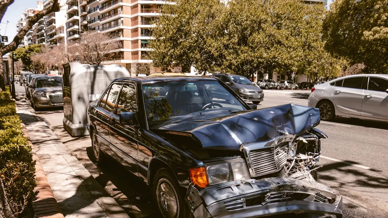 An older sedan with a crushed front end parked on a city street; badges and plates blurred.